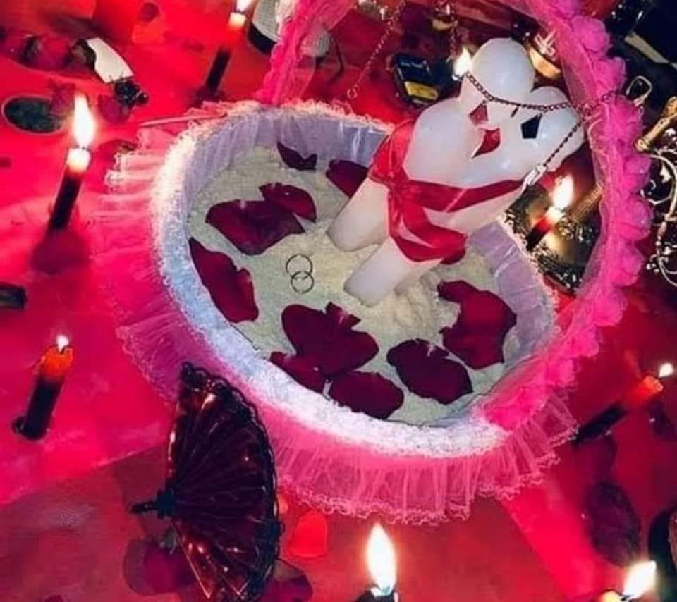 Trabalho de amarração com vela em formato de corpo sobre pétalas de rosa vermelhas, duas alianças e velas acesas ao redor