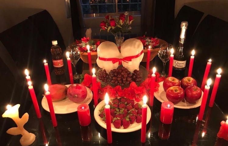 Mesa de trabalho com velas vermelhas acesas, morangos, maçãs, uvas, champagne e duas figuras unidas por uma fita vermelha